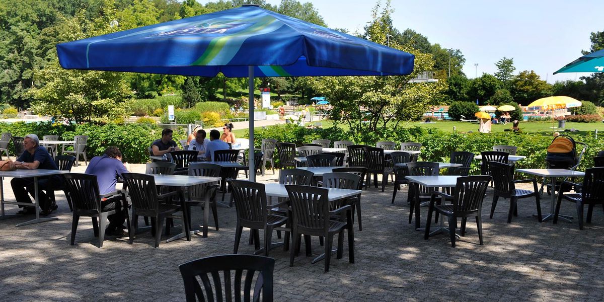 Leeres Aussencafé mit weissen Tischen und schwarzen Stühlen unter blauen Sonnenschirmen, umgeben von grüner Landschaft.