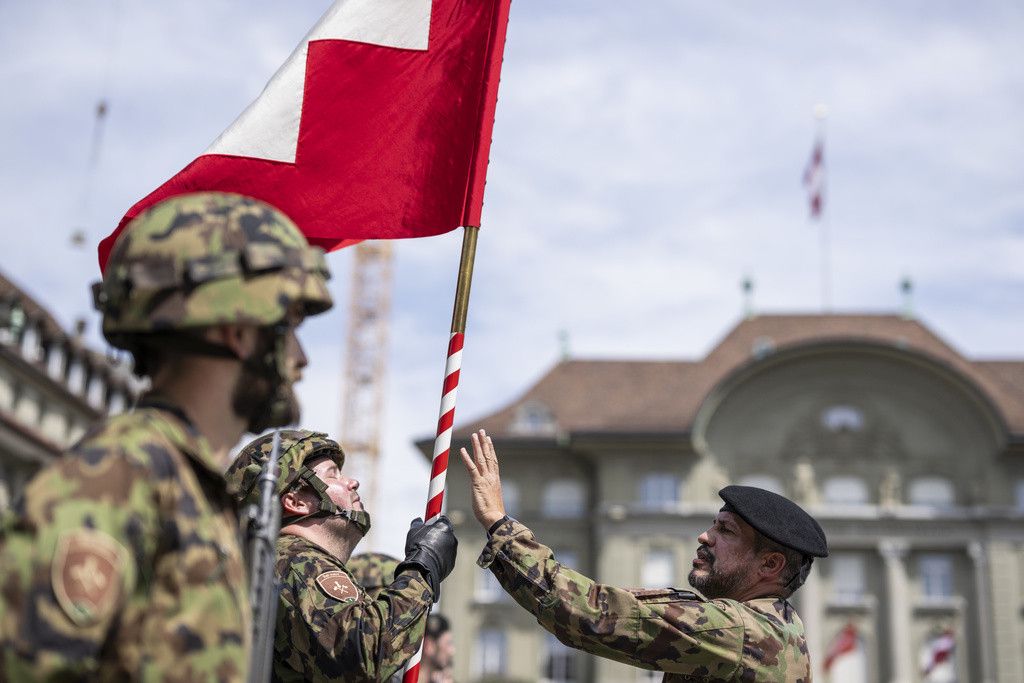 Des soldats suisses en uniforme tiennent le drapeau suisse lors d'une cérémonie devant un bâtiment officiel avec des drapeaux.