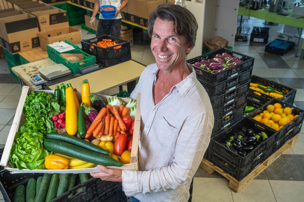 Michael Dusong, le fondateur de l'entreprise UglyFruit, tenant un panier de fruits et légumes bio variés, dans un entrepôt avec des caisses de produits en arrière-plan.