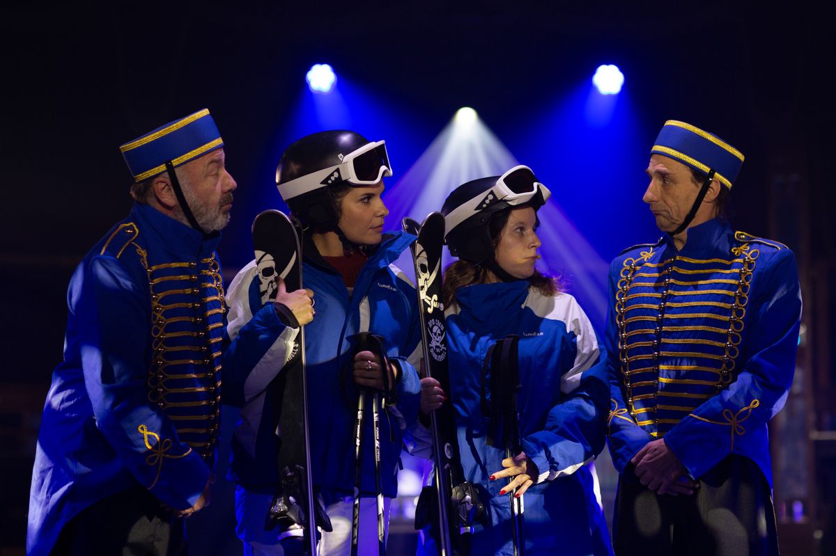 Quatre personnes en uniforme bleu et or avec des skis sous des lumières de scène.