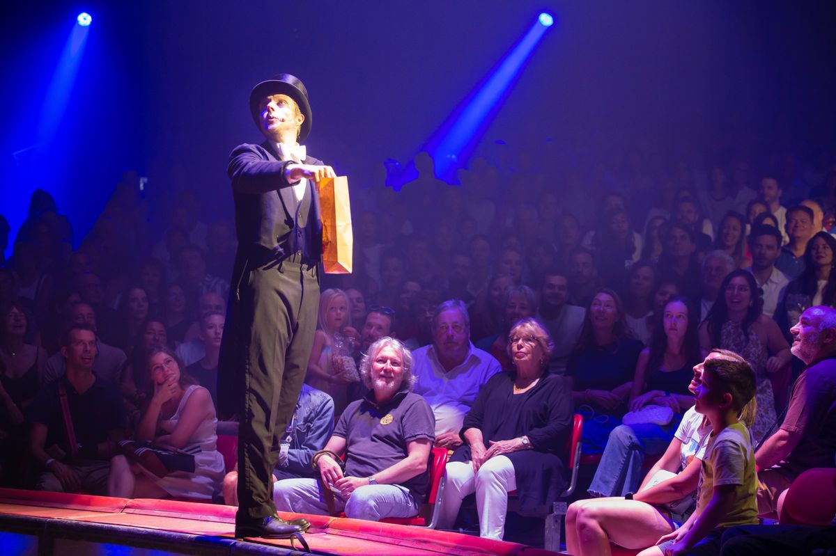 Un magicien réalise un tour de magie sur scène avec des spectateurs assis autour, éclairés par des projecteurs bleus.