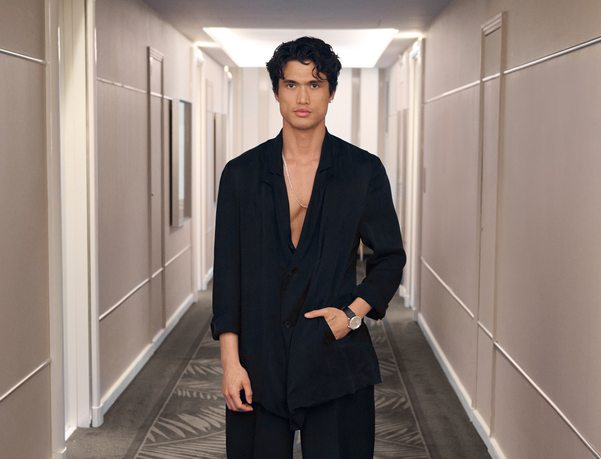 A young man in a smart black suit, open collar and watch on his wrist, posing in a bright hotel corridor.