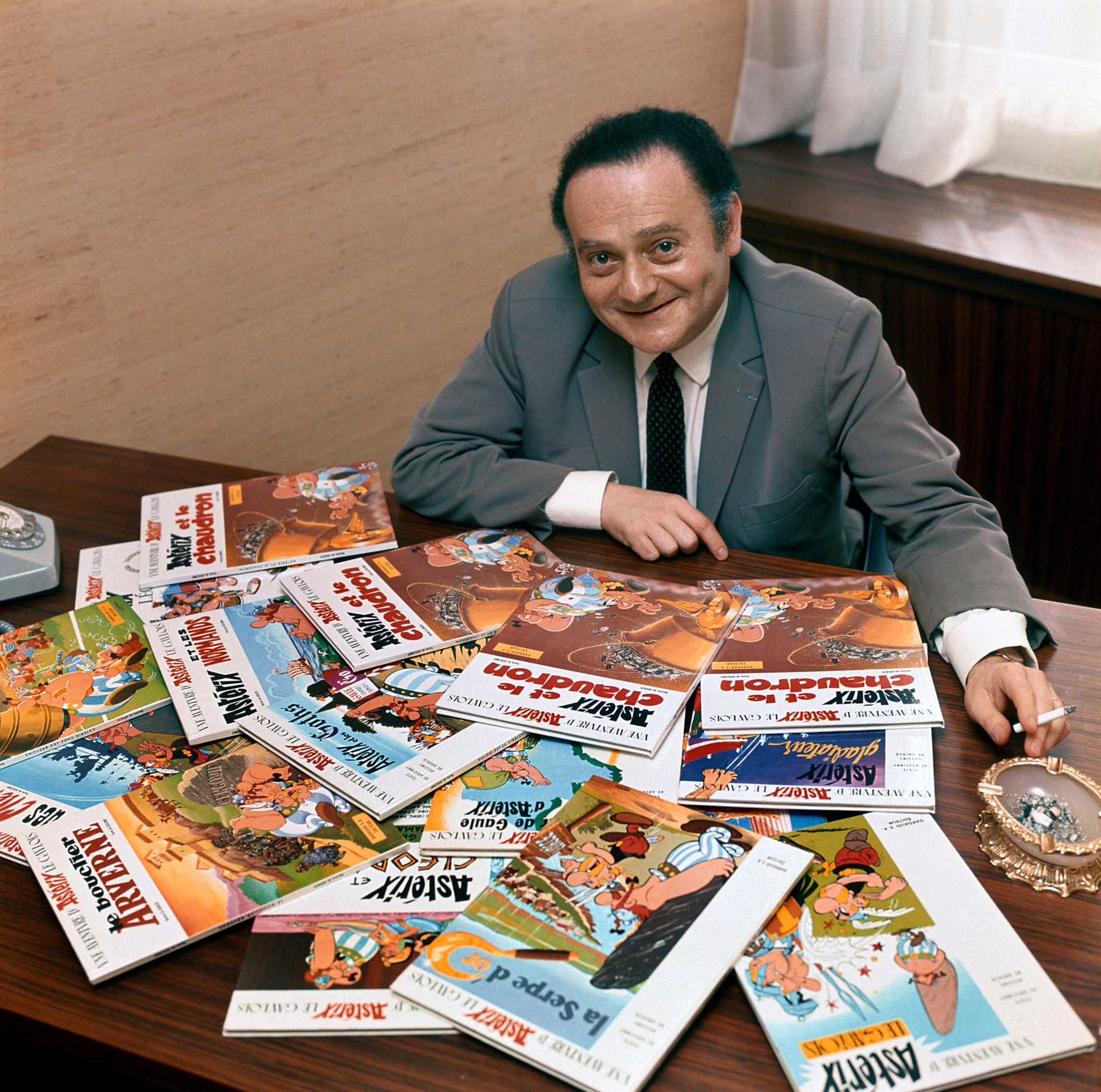 René Goscinny, Autor von Asterix, Lucky Luke und Isnogud, posiert um 1969 lächelnd hinter einem Tisch voller Asterix-Comichefte.