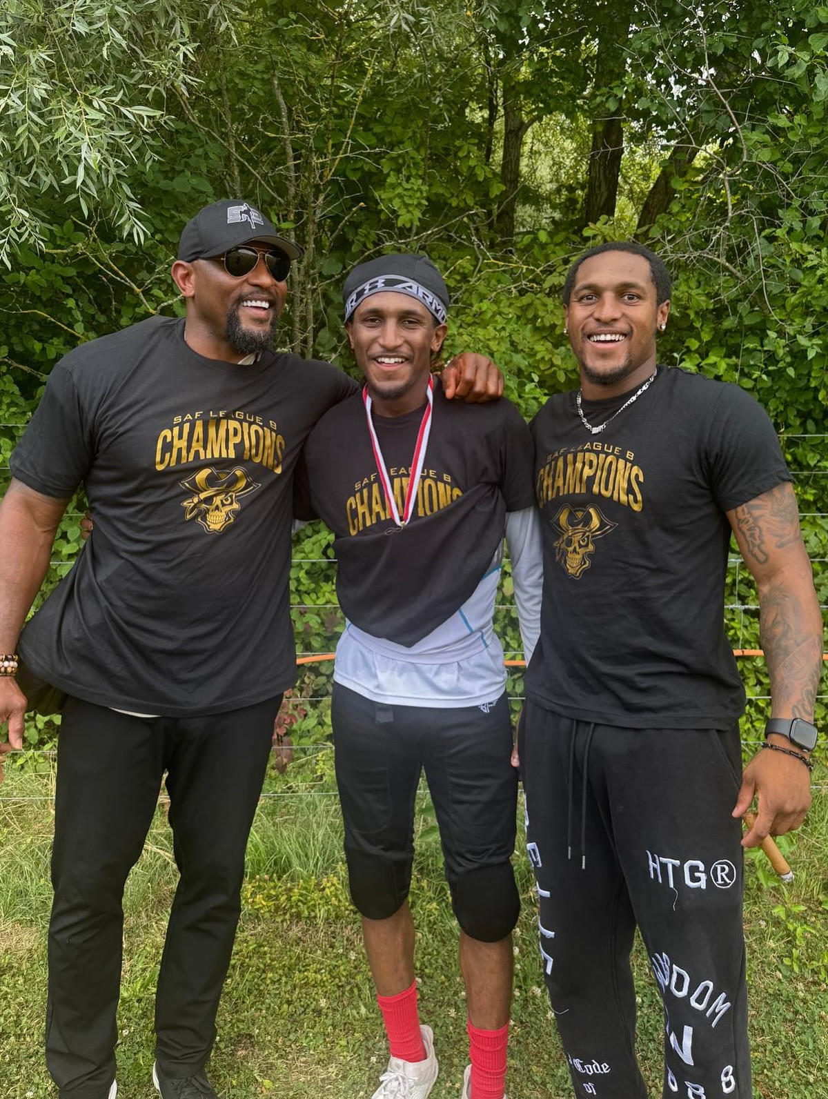 Drei Männer in schwarzen T-Shirts mit ’Champions’-Aufdruck stehen lächelnd im Freien zusammen.