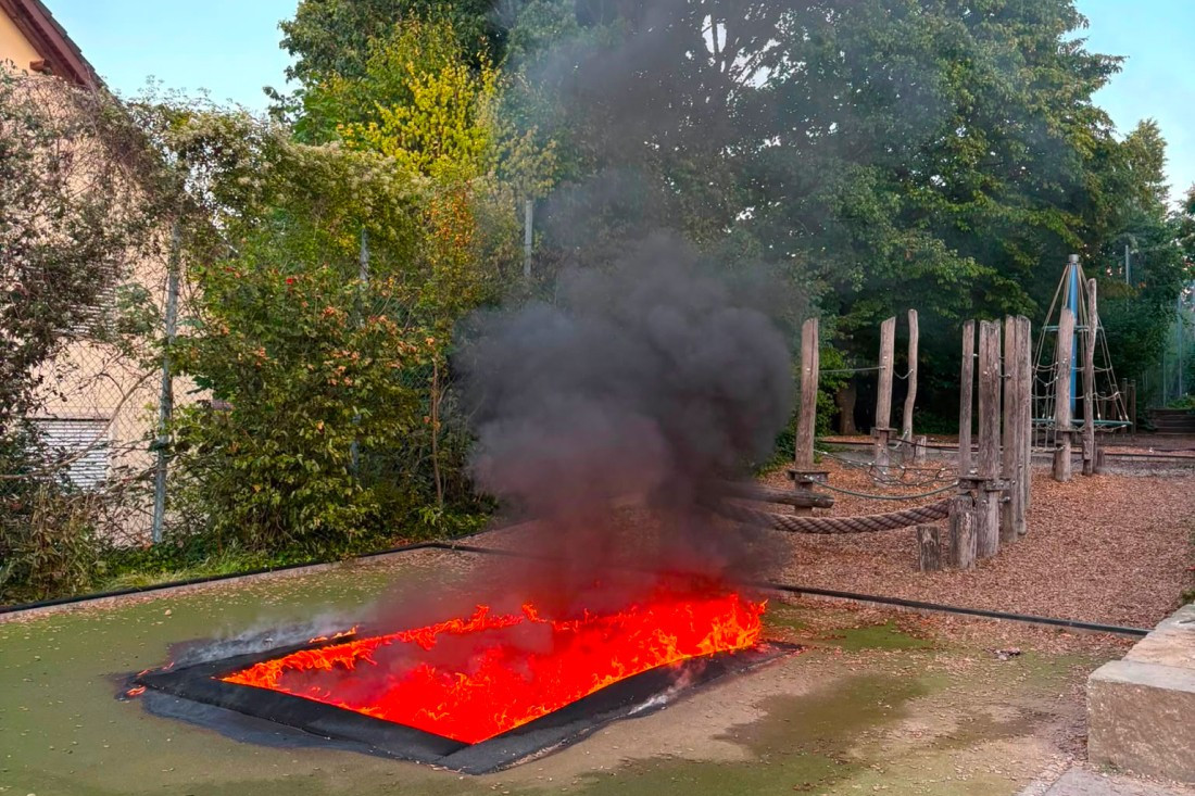 Brennendes Trampolin auf einem Spielplatz mit dichtem schwarzen Rauch, umgeben von Bäumen und Klettergerüsten.