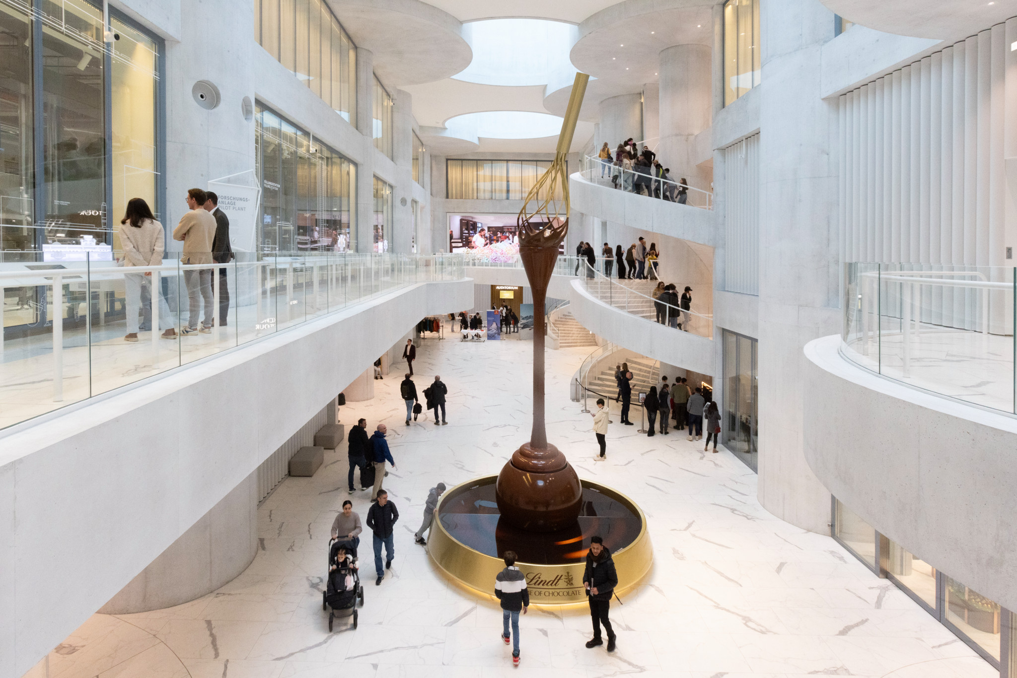 Imposante Schokoladenfontäne im Atrium des Lindt Home of Chocolate Museums in Kilchberg, umgeben von Besuchern auf mehreren Ebenen.