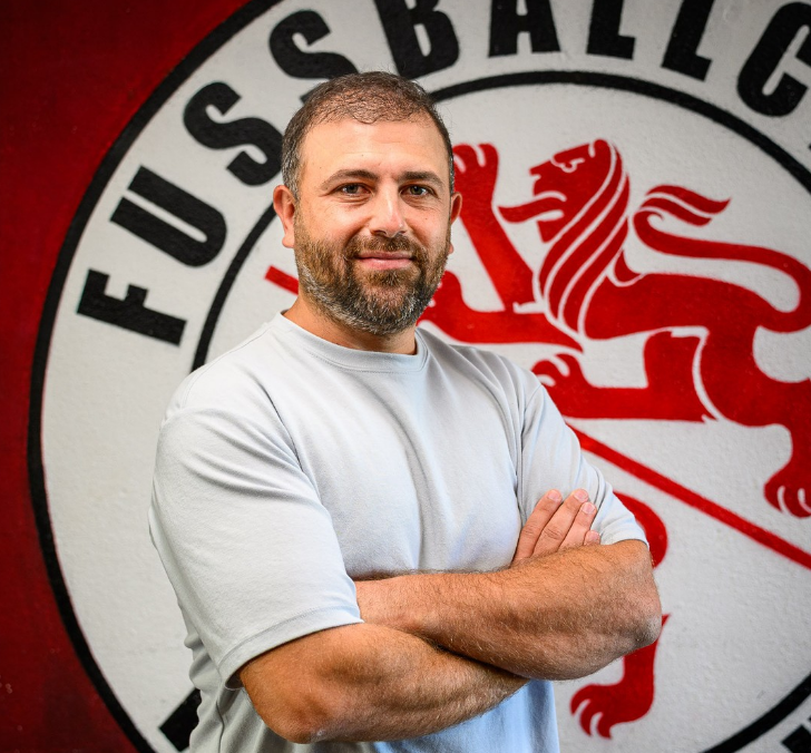 Mann mit verschränkten Armen vor rot-weissem Fussballclub-Logo mit Löwenwappen
