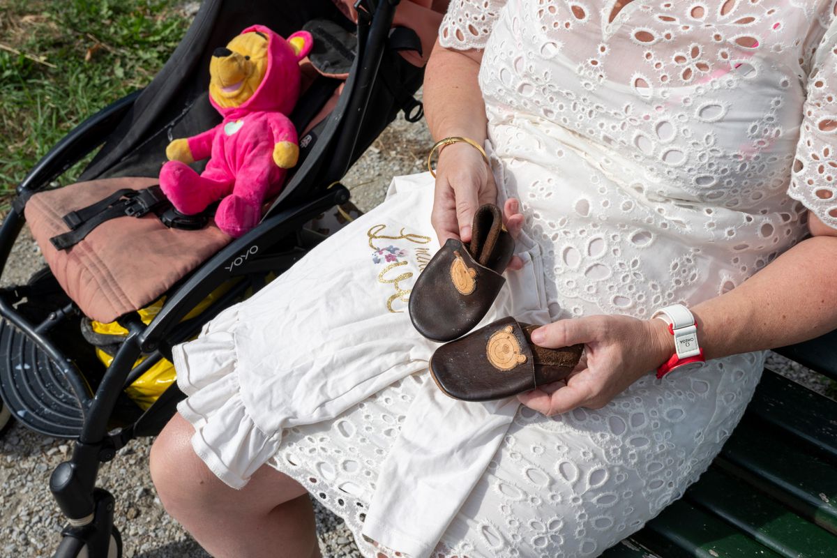 Une femme en robe blanche assise sur un banc tenant des chaussures pour enfant à côté d'une poussette avec une peluche rose. ©Florian Cella/24H