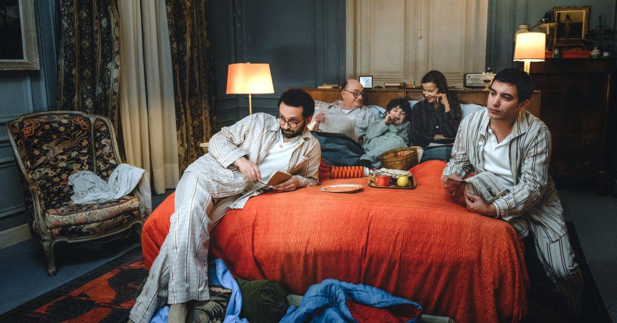 Cinq personnes en pyjama, détendues dans une chambre sur un grand lit recouvert d’une couverture rouge, entourées de coussins et d’objets divers.