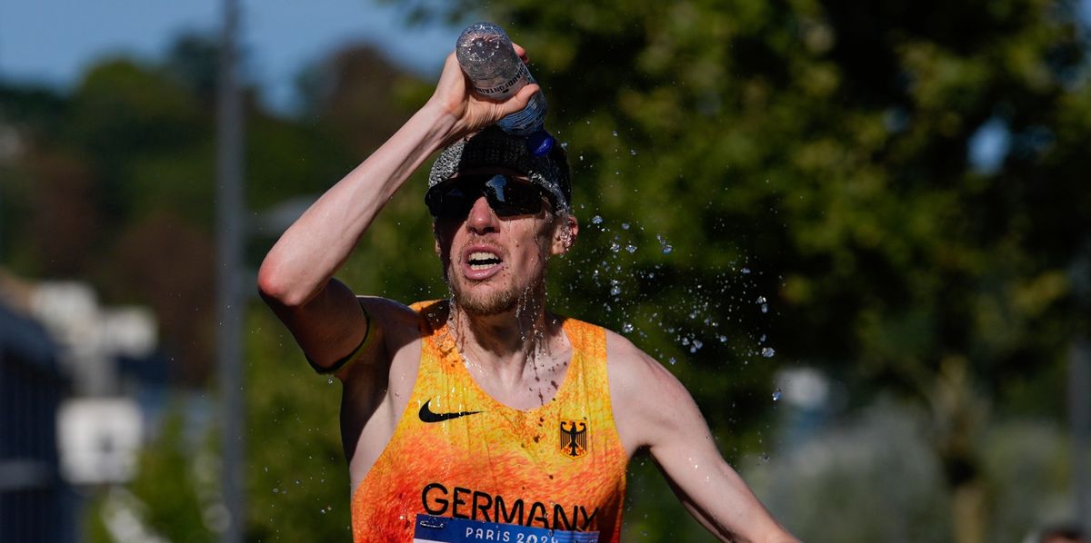 Un coureur allemand verse de l'eau sur sa tête lors d'une course, portant un maillot orange avec l'inscription 'GERMANY' et 'RINGER', et des lunettes de soleil.