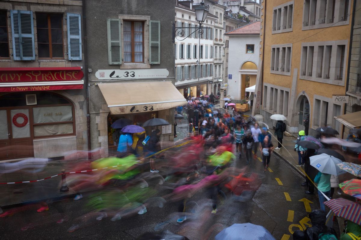 Coureurs sous la pluie lors de la 46e course de l’Escalade à Genève, traversant des rues bordées d’immeubles anciens.
