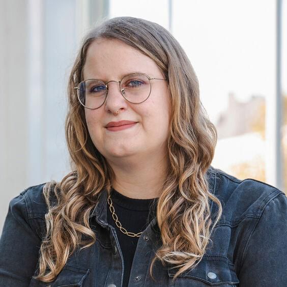 Femme avec des lunettes et une veste en jean, souriant légèrement devant une fenêtre.