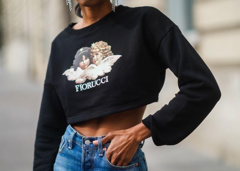 Personne portant un sweat court noir avec une image d’anges et le texte ’Fiorucci’, tenant leur taille avec une main, en jean.