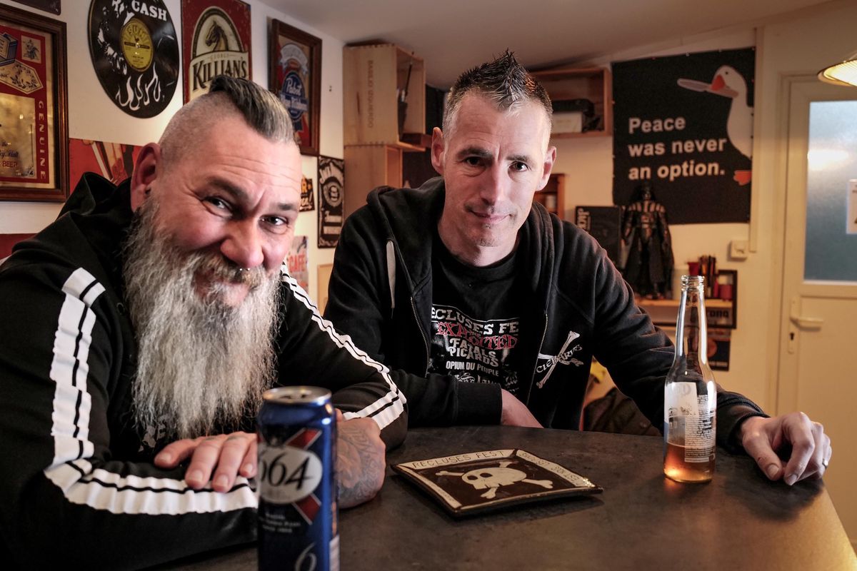 Deux organisateurs du Recluses Fest à Genève posent dans un bar, entourés de décorations punk, tenant des boissons.