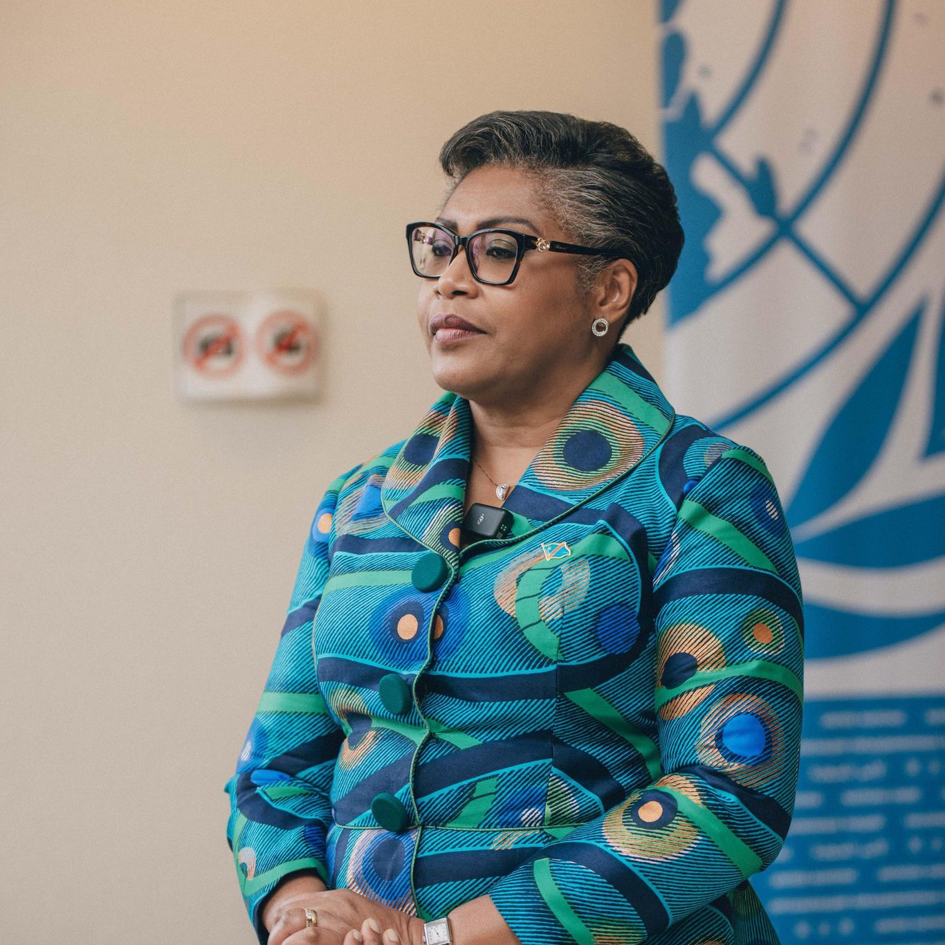 Une femme portant des lunettes et une tenue colorée se tient debout, de profil, devant un panneau avec des motifs géométriques bleus.
