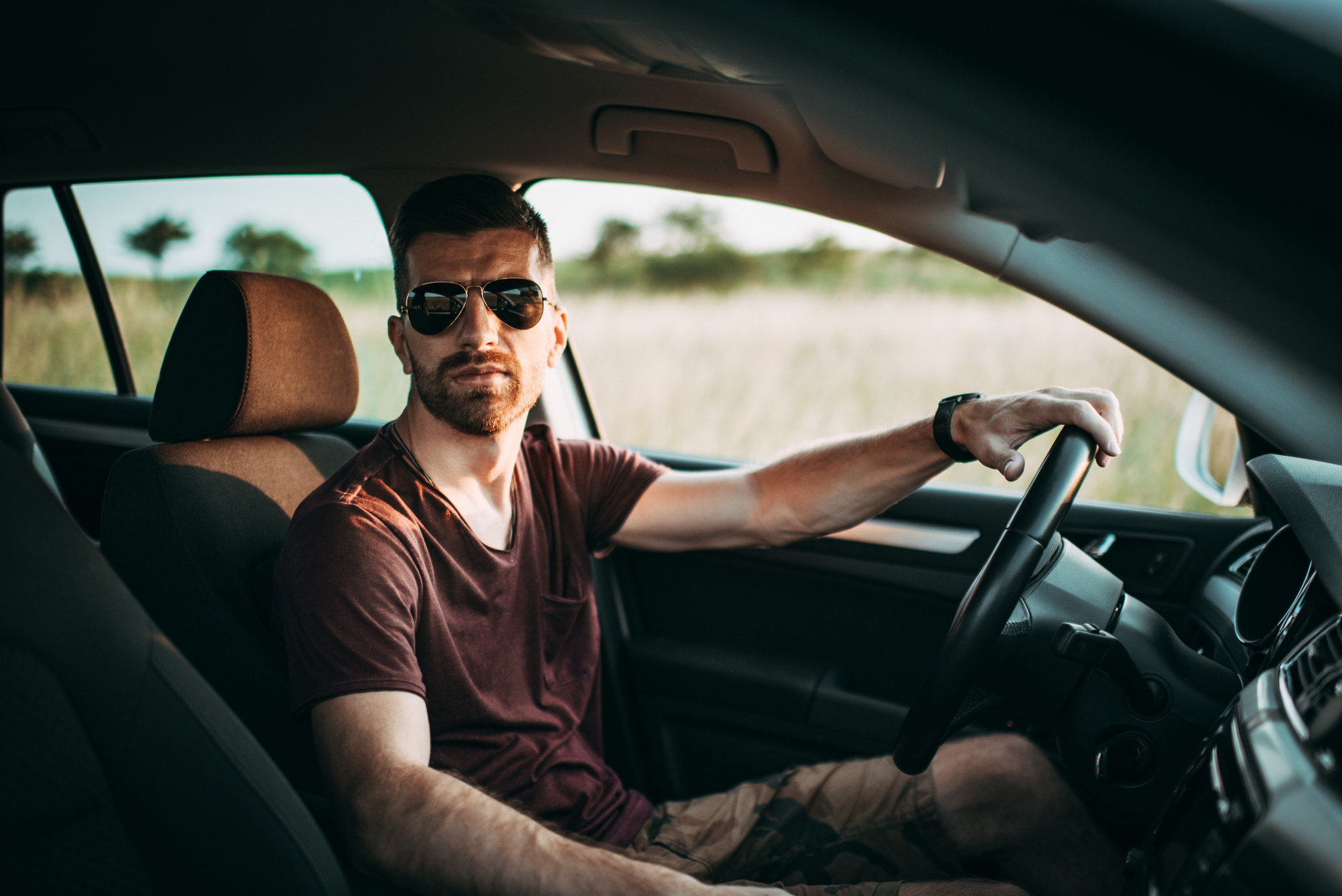Junger Mann mit Sonnenbrille und rotem T-Shirt sitzt am Steuer eines Autos auf einer Landstrasse.