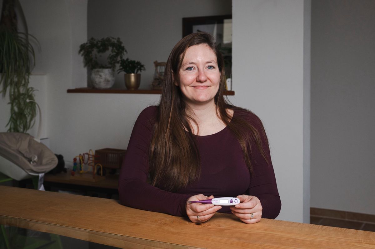 Une femme aux longs cheveux bruns, tenant un thermomètre, est appuyée sur un comptoir en bois dans un intérieur décoré de plantes. Elle sourit légèrement.