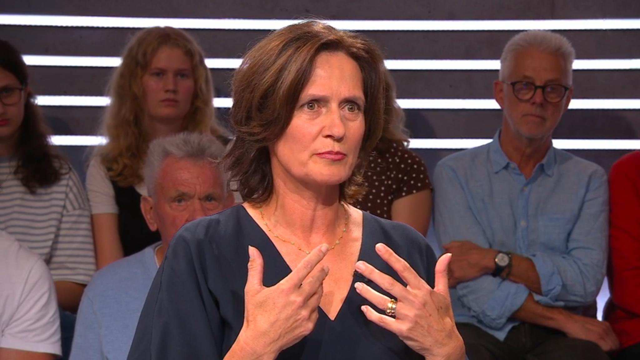 Frau in blauem Oberteil gestikuliert beim Sprechen in einer Talkshow, Publikum im Hintergrund.