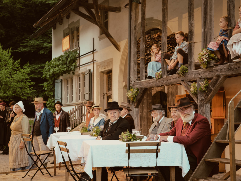 Aussenaufnahme einer Theateraufführung von ’Geltstag’ nach Jeremias Gotthelf im Landschaftstheater Ballenberg, mit Darstellern in historischen Kostümen an langen Tischen.