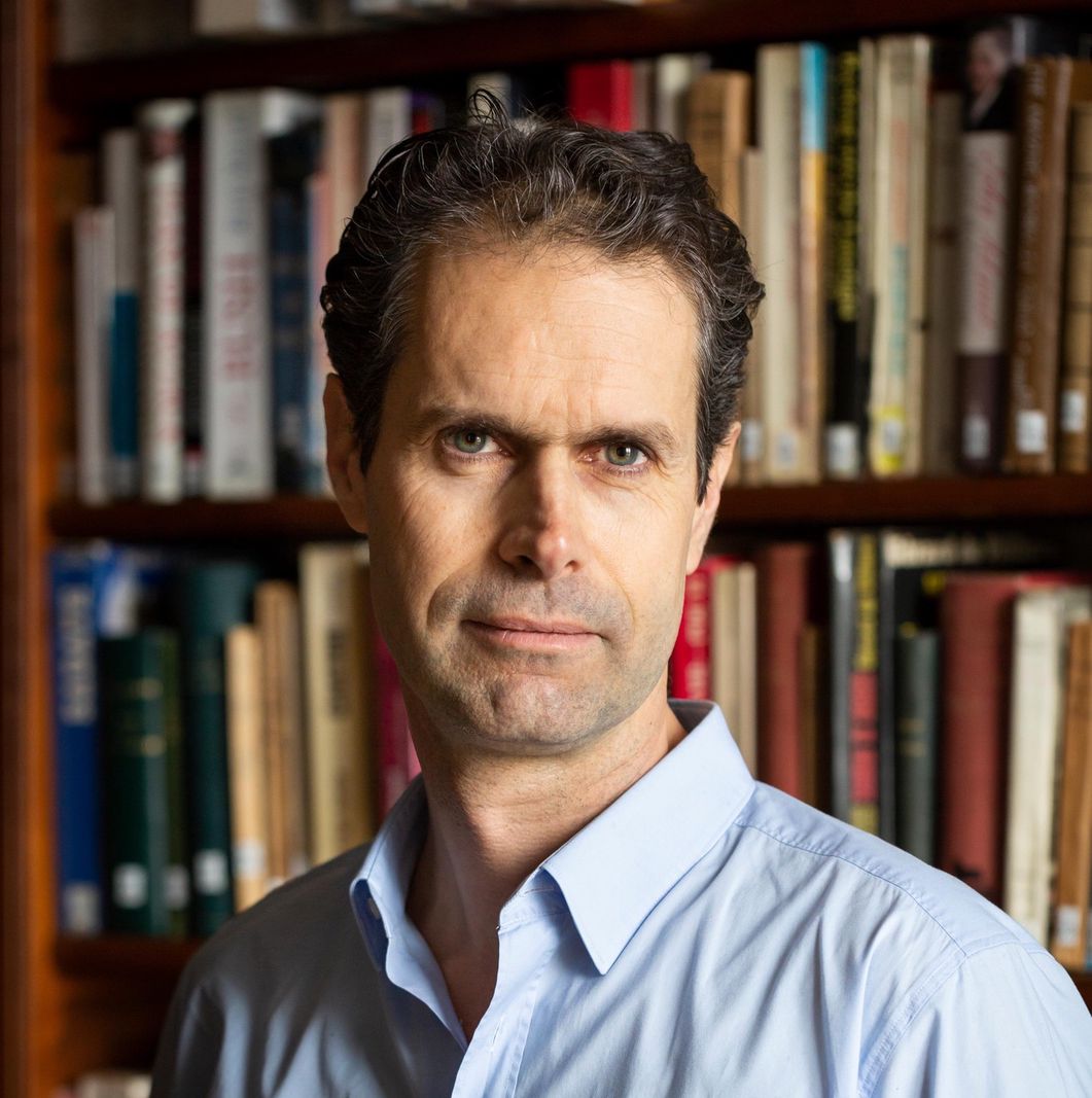 Homme en chemise bleue debout devant une étagère remplie de livres.