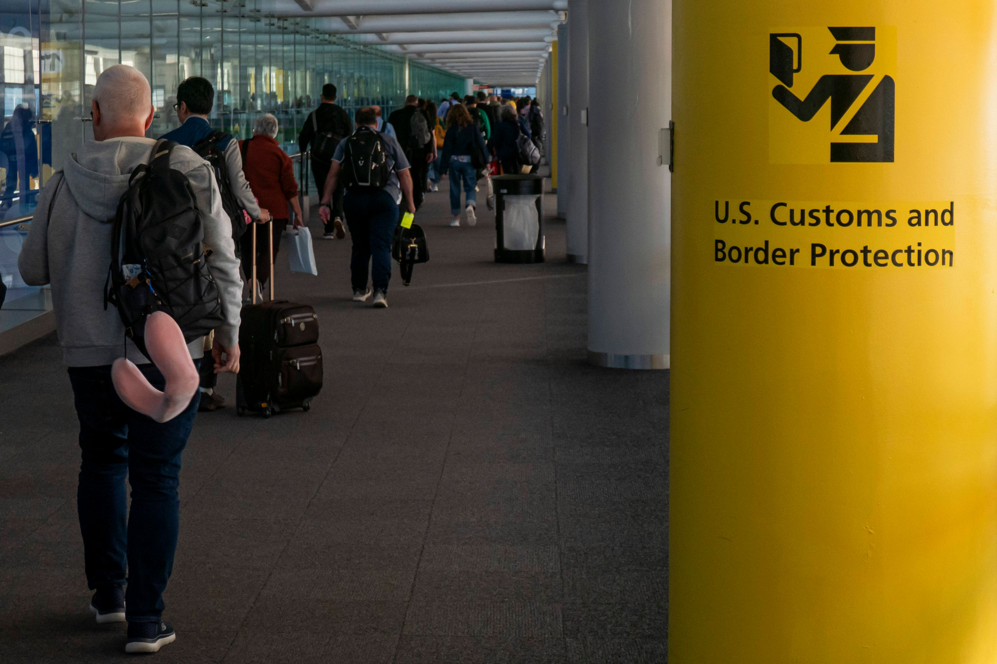 Passagiere gehen im Terminal des Newark Liberty International Airport in Richtung Passkontrolle, neben einem Pfeiler mit dem Symbol und der Aufschrift der U.S. Customs and Border Protection auf gelbem Hintergrund.