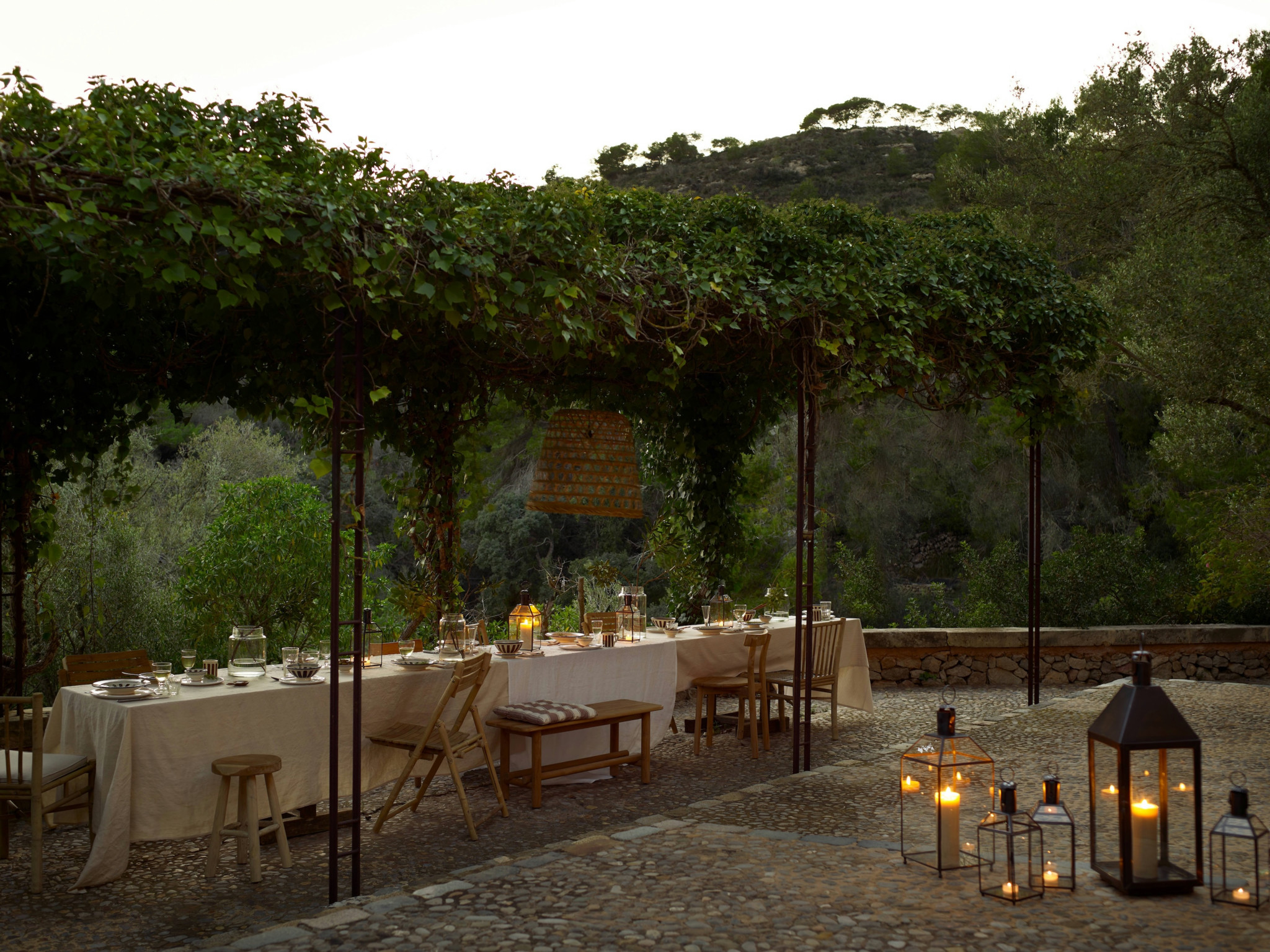 Gedeckter Tisch unter begrünter Pergola im Freien, beleuchtet von Laternen in mediterraner Abendatmosphäre.