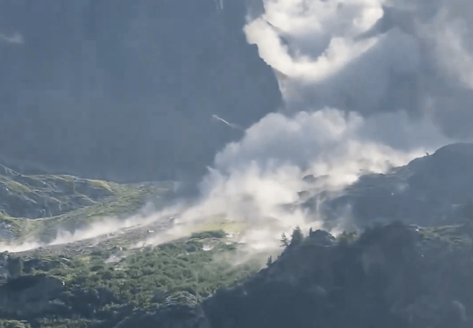 Un glissement de terrain dans une région montagneuse, avec des nuages de poussière et de débris visibles sur les pentes verdoyantes.