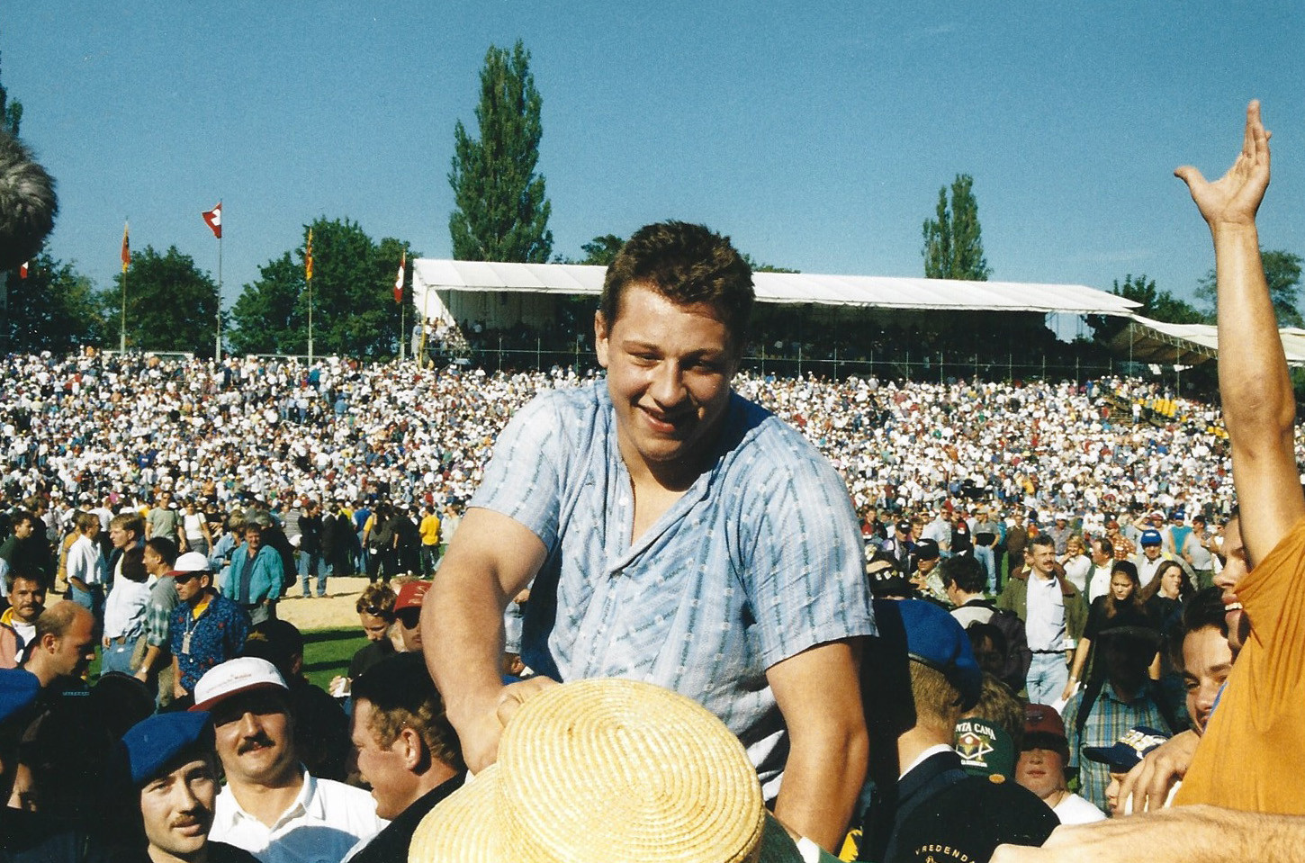 Junger Mann im blauen Hemd lächelt inmitten einer grossen Menschenmenge bei einer Freiluftveranstaltung, mit Tribünen im Hintergrund. Junger Mann im blauen Hemd lächelt inmitten einer grossen Menschenmenge bei einer Freiluftveranstaltung, mit Tribünen im Hintergrund.