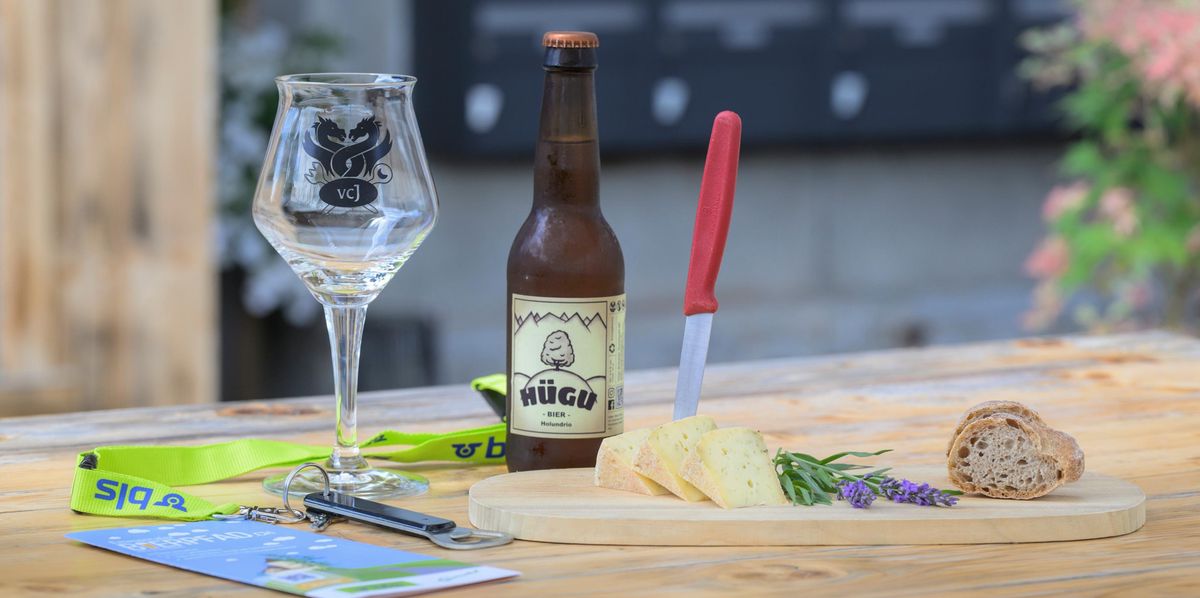 Eine Flasche Hügu-Bier steht auf einem Holztisch, daneben ein leeres Glas und eine Aperoplatte.