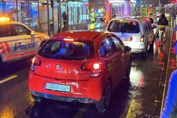 Scène de nuit pluvieuse en ville avec des voitures alignées, dont une voiture rouge au premier plan, et des véhicules de police et de secours visibles en arrière-plan.
