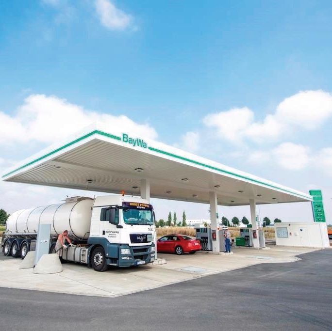 Tankstelle der Marke BayWa mit Lkw und rotem Auto unter einem überdachten Bereich, umgeben von ländlicher Landschaft.