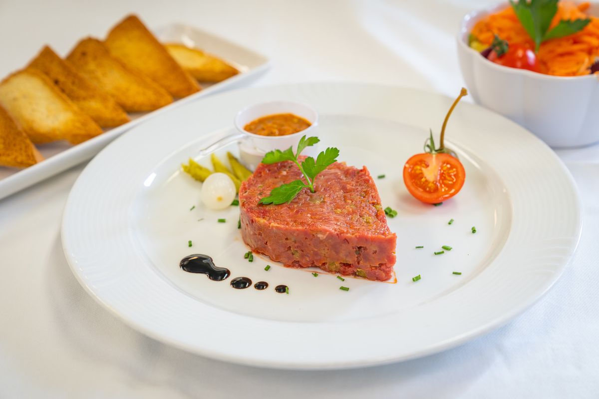 Un plat de tartare de bœuf servi avec des toasts, une tomate cerise, des cornichons et une petite sauce.