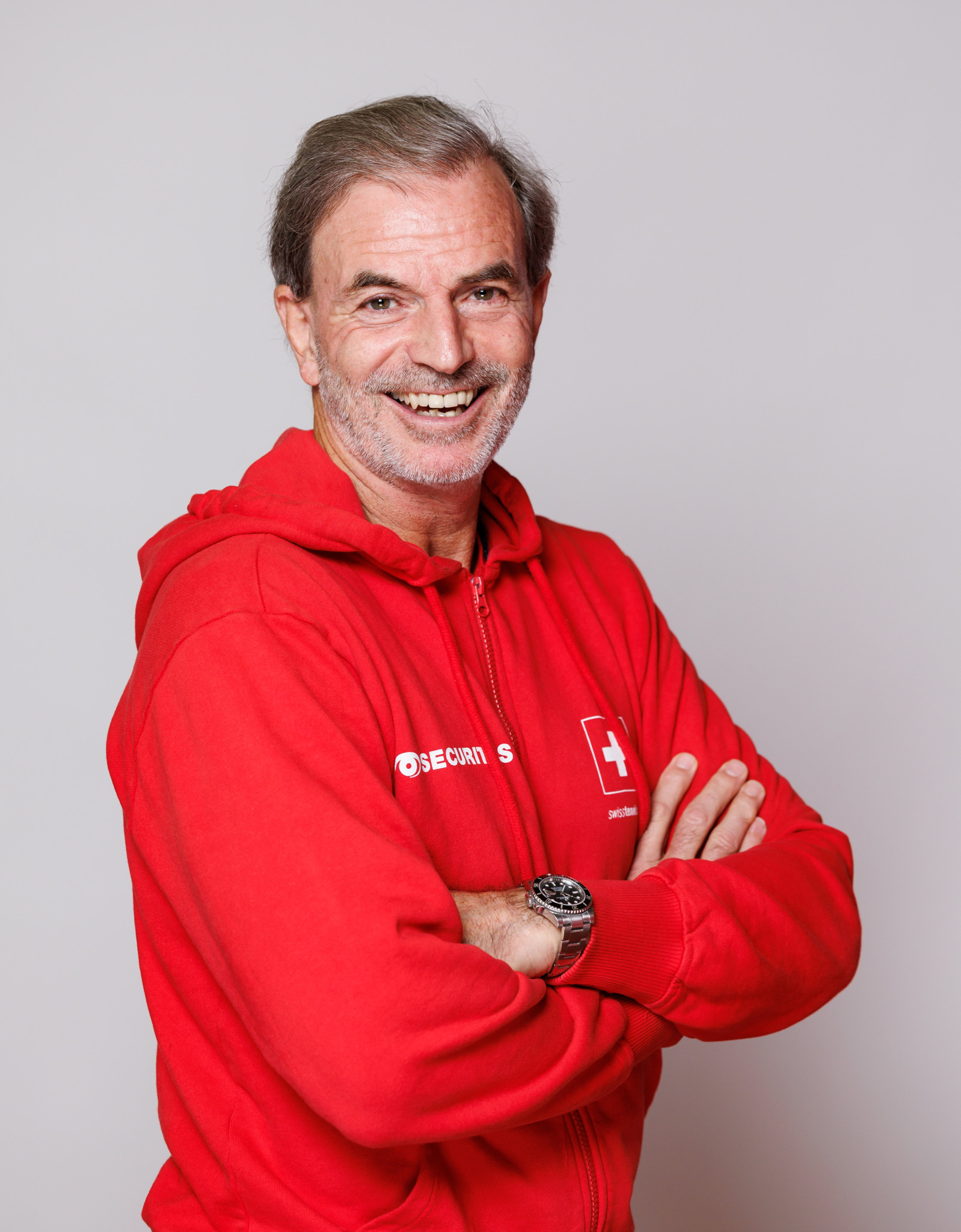 Heinz Guenthardt, Kapitän des Schweizer Teams, in rotem Hoodie vor neutralem Hintergrund, posiert lächelnd mit verschränkten Armen.