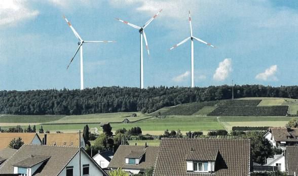 Drei Windkraftanlagen vor einem bewaldeten Hügel, im Vordergrund mehrere Wohnhäuser.