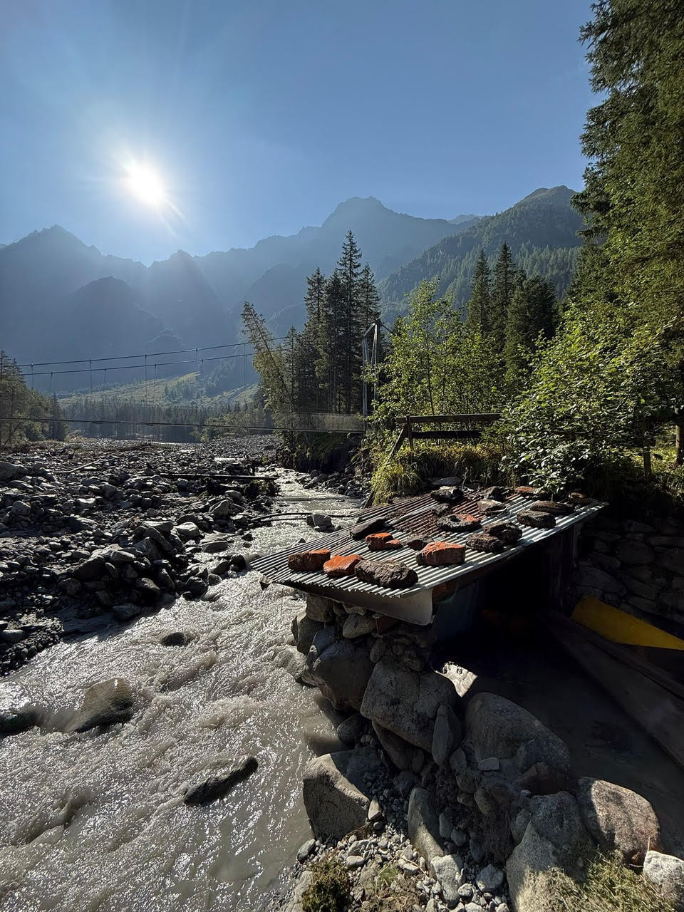 Sonnenbeschienene Berglandschaft mit einem reissenden Fluss, einer kleinen Holzhütte am Ufer und einem Hängebrücke im Hintergrund.