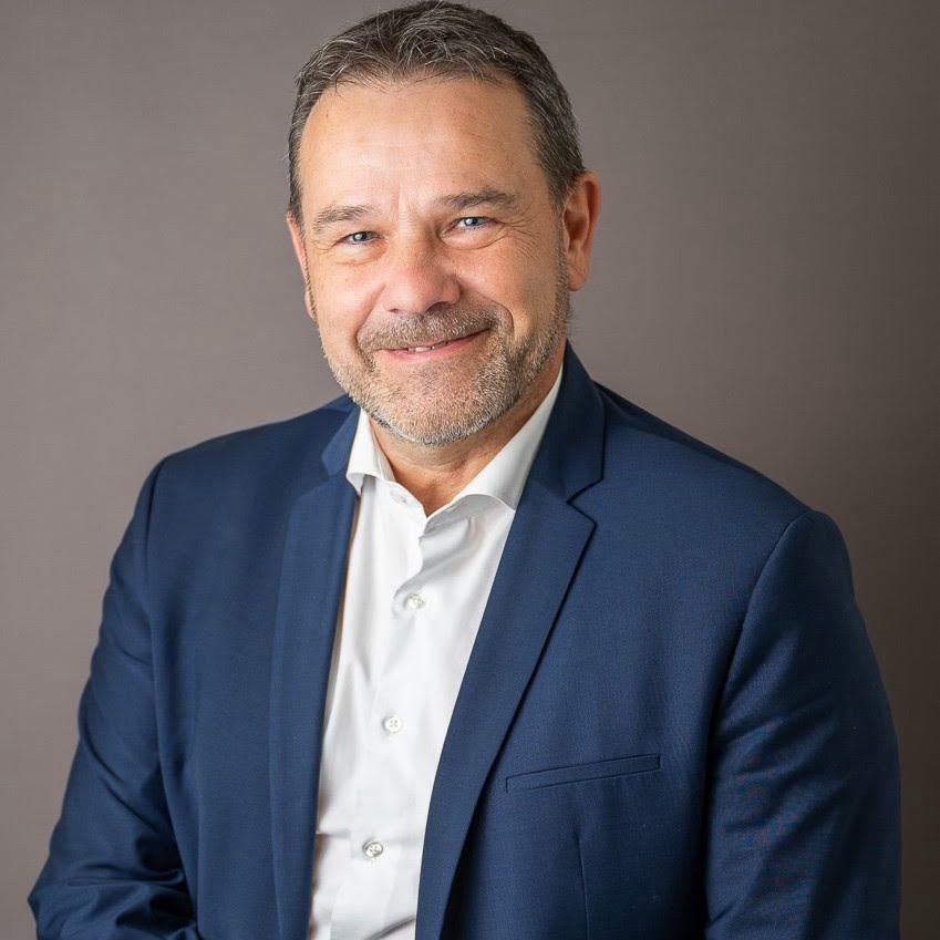 Homme souriant en costume bleu et chemise blanche posant devant un fond gris.