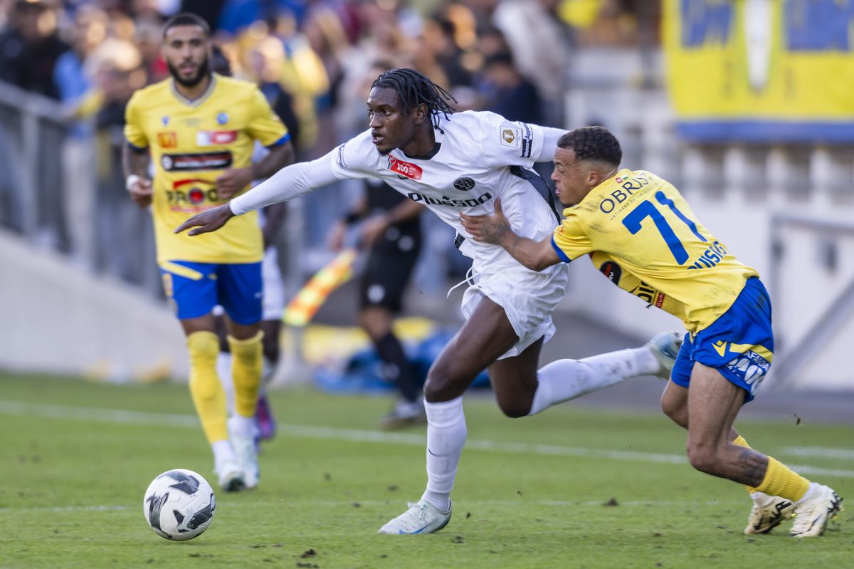 Un joueur de football en blanc court avec le ballon devant lui, poursuivi par un joueur en jaune et bleu, tandis qu'un autre joueur en jaune regarde la scène.