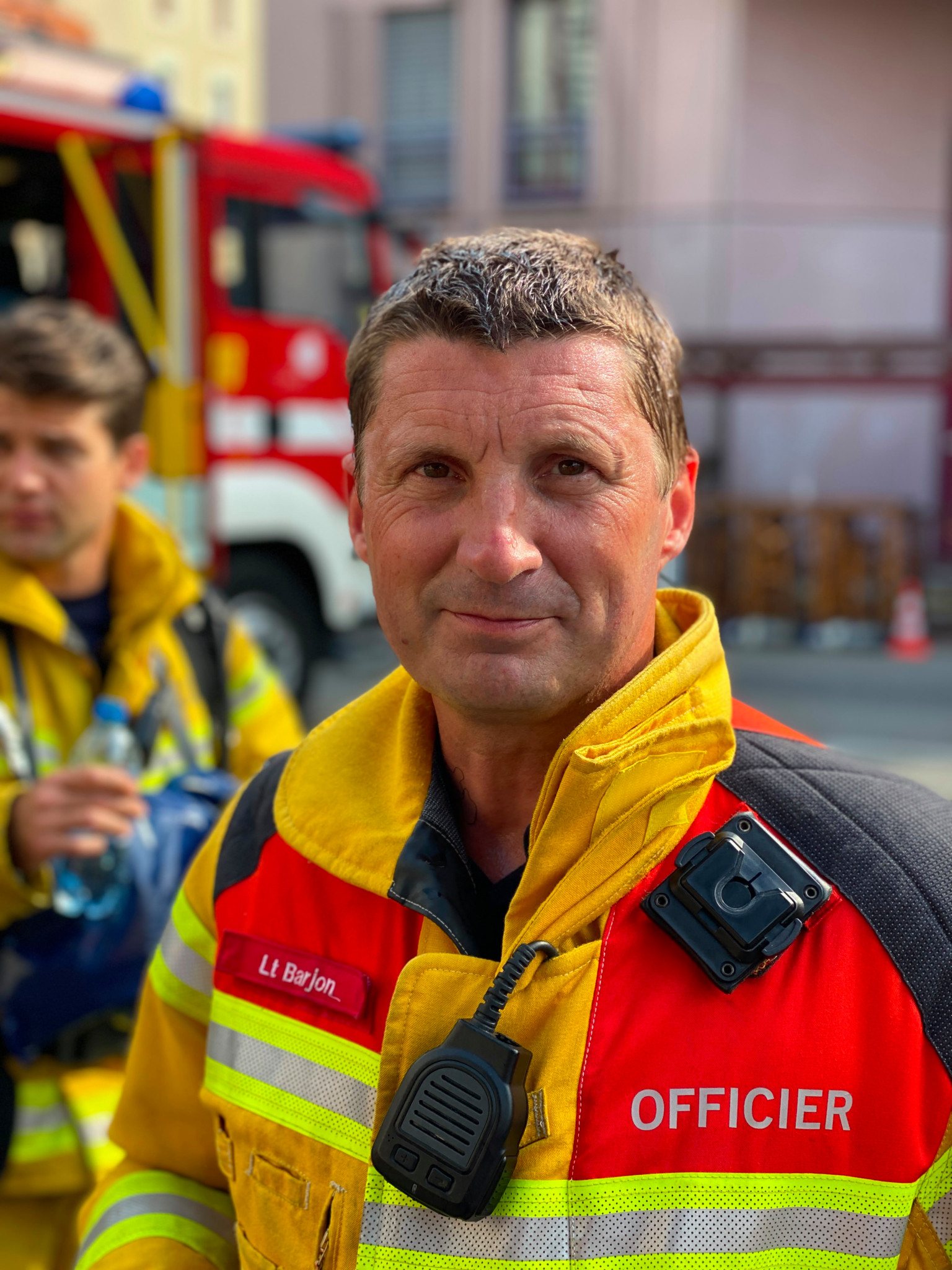 Un officier des pompiers, Lt Birjon, en tenue jaune et rouge avec radio, devant un camion de pompiers rouge. Un officier des pompiers, Lt Birjon, en tenue jaune et rouge avec radio, devant un camion de pompiers rouge.