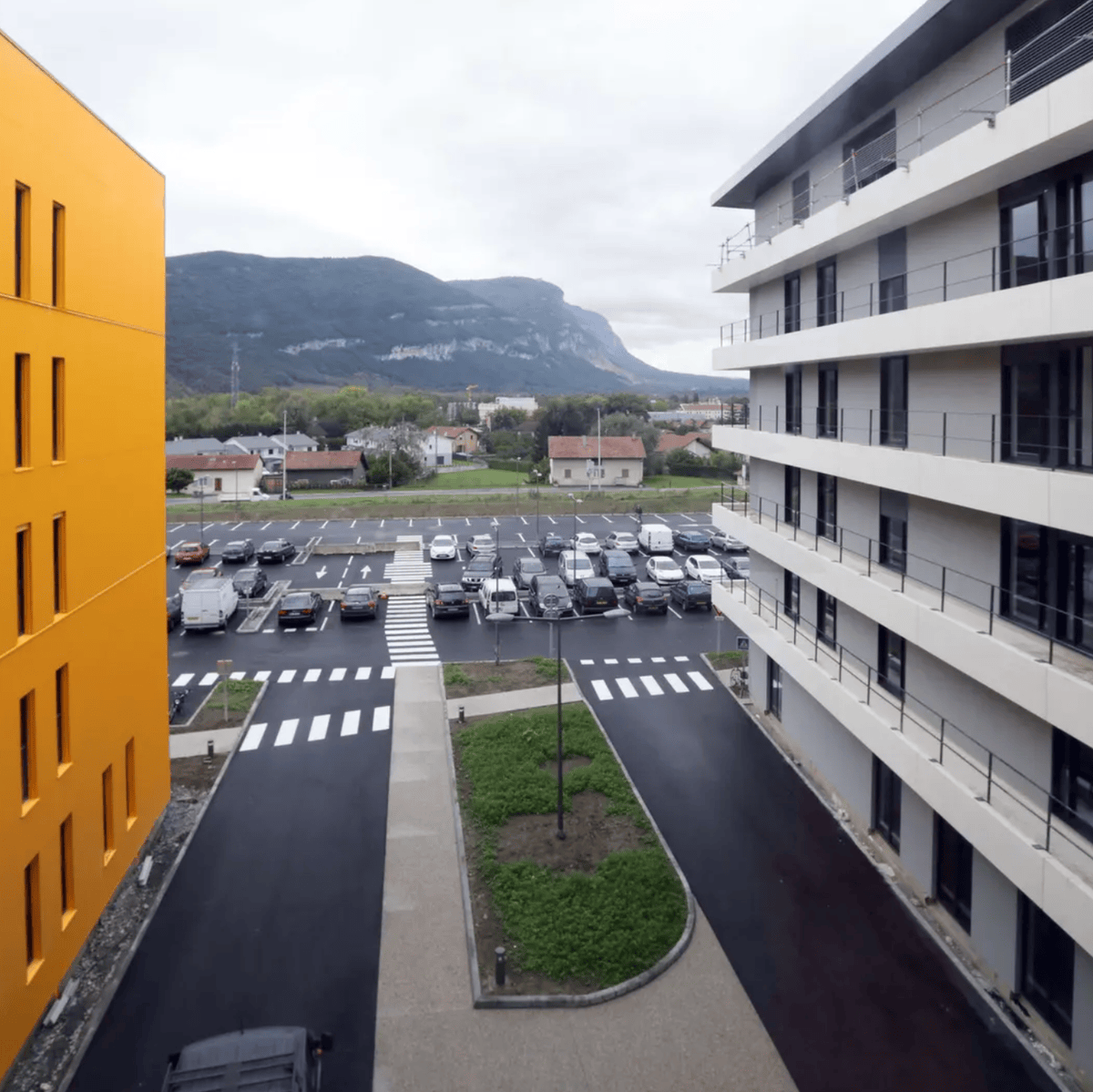 Vue de deux immeubles modernes encadrant un parking, avec des montagnes en arrière-plan.