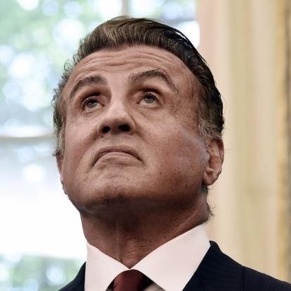 Un homme en costume regarde vers le haut dans une pièce avec des rideaux dorés et une fenêtre en arrière-plan.