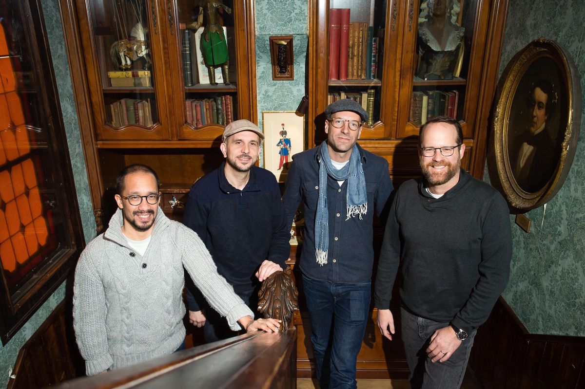 L’équipe de l’escape game Grand Trip Trap Hôtel à Carouge, comprenant Khoa Hoang, Nicolas Aubry, Aurèle Barde et Gian Cla Pinösh, posant dans un intérieur boisé élégant avec des livres et des objets décoratifs en arrière-plan.