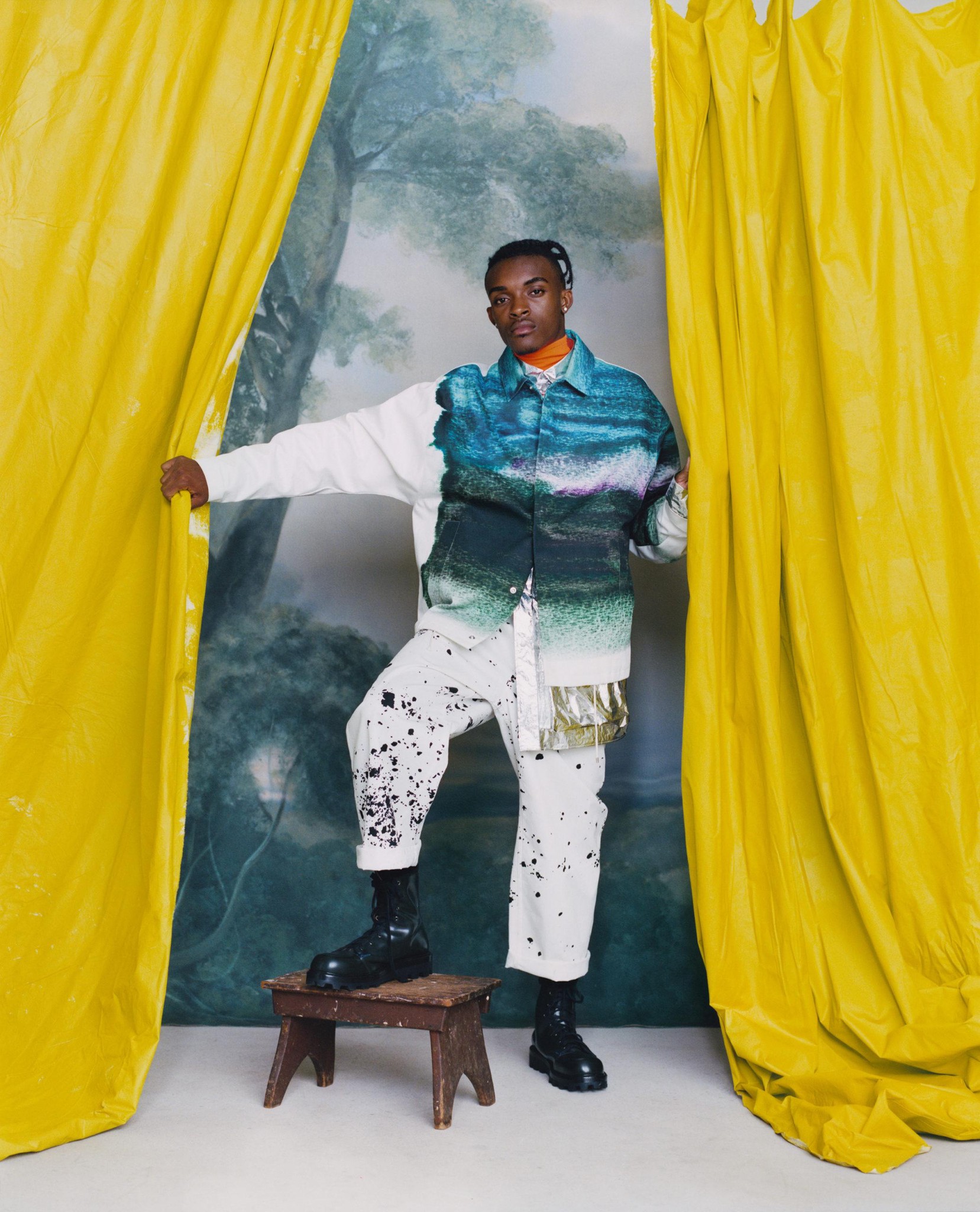 Personne posant entre des rideaux jaunes avec un fond de paysage, portant des vêtements aux couleurs vibrantes et des bottes noires.