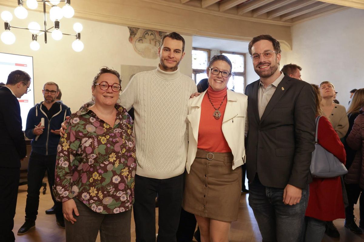 Groupe de quatre personnes souriantes dans une pièce éclairée, entourées de plusieurs autres personnes en arrière-plan.