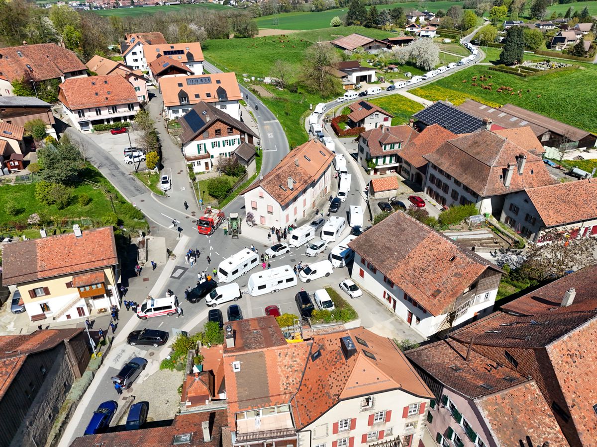 Vue aérienne d'un village suisse avec des maisons aux toits rouges et des chemins sinueux. Plusieurs camping-cars sont stationnés dans le village.