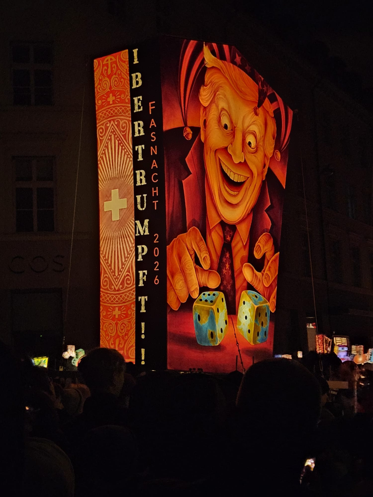 Installation de lanternes lors d'un défilé nocturne avec représentation d'un homme souriant tenant des dés. Le texte dit « IBER TRUMP !! CARNAVAL 2026'.