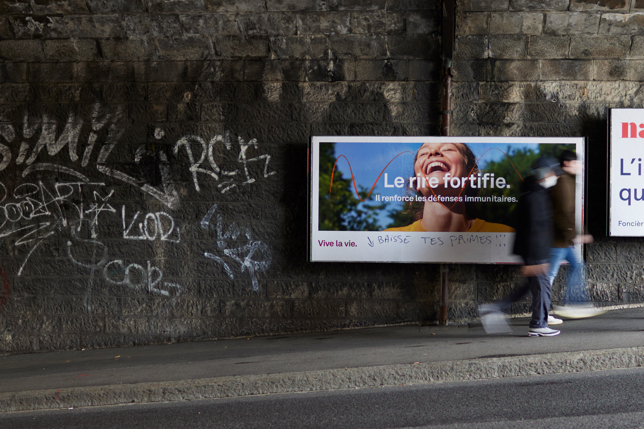 Un panneau publicitaire sous un pont, avec des graffitis sur le mur à côté. L'affiche publicitaire montre une femme souriante avec le texte 'Le rire fortifie. Il renforce les défenses immunitaires.' Deux personnes passent devant l'affiche.