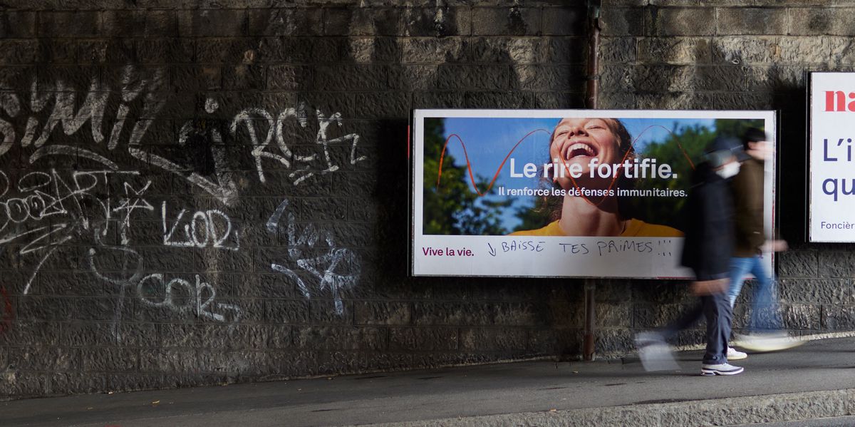Un panneau publicitaire sous un pont, avec des graffitis sur le mur à côté. L'affiche publicitaire montre une femme souriante avec le texte 'Le rire fortifie. Il renforce les défenses immunitaires.' Deux personnes passent devant l'affiche.