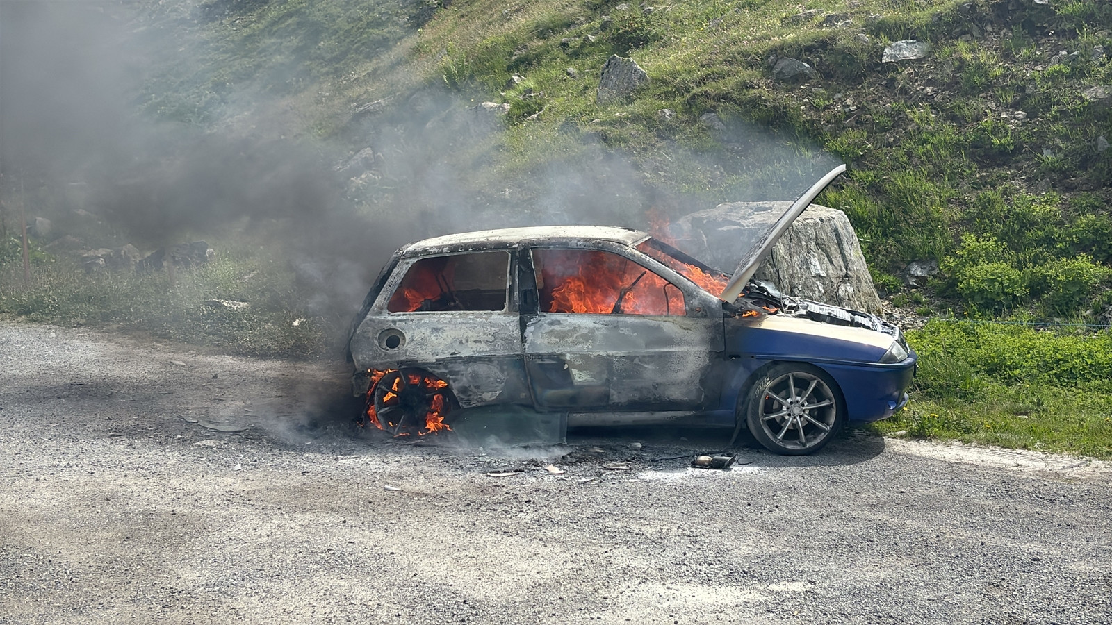Feuerwehrfahrzeuge und Einsatzkräfte auf einer Alpenpassstrasse mit aufsteigendem Rauch, umgeben von grünen Bergwiesen. Feuerwehrfahrzeuge und Einsatzkräfte auf einer Alpenpassstrasse mit aufsteigendem Rauch, umgeben von grünen Bergwiesen.