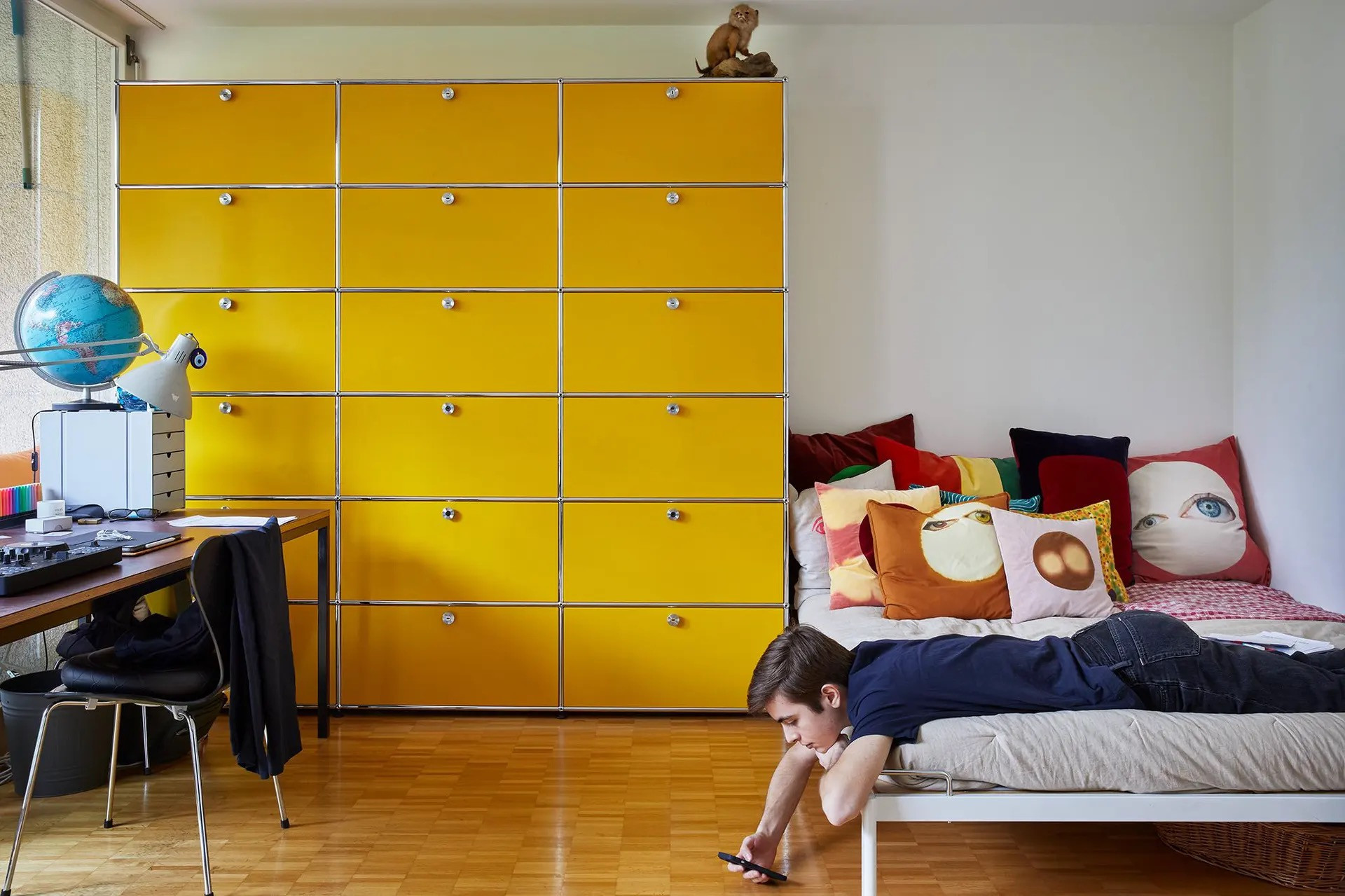 Junge liegt auf einem Bett mit bunten Kissen in einem Kinderzimmer mit auffälligem gelbem Schranksystem und einem Schreibtisch mit Globus.