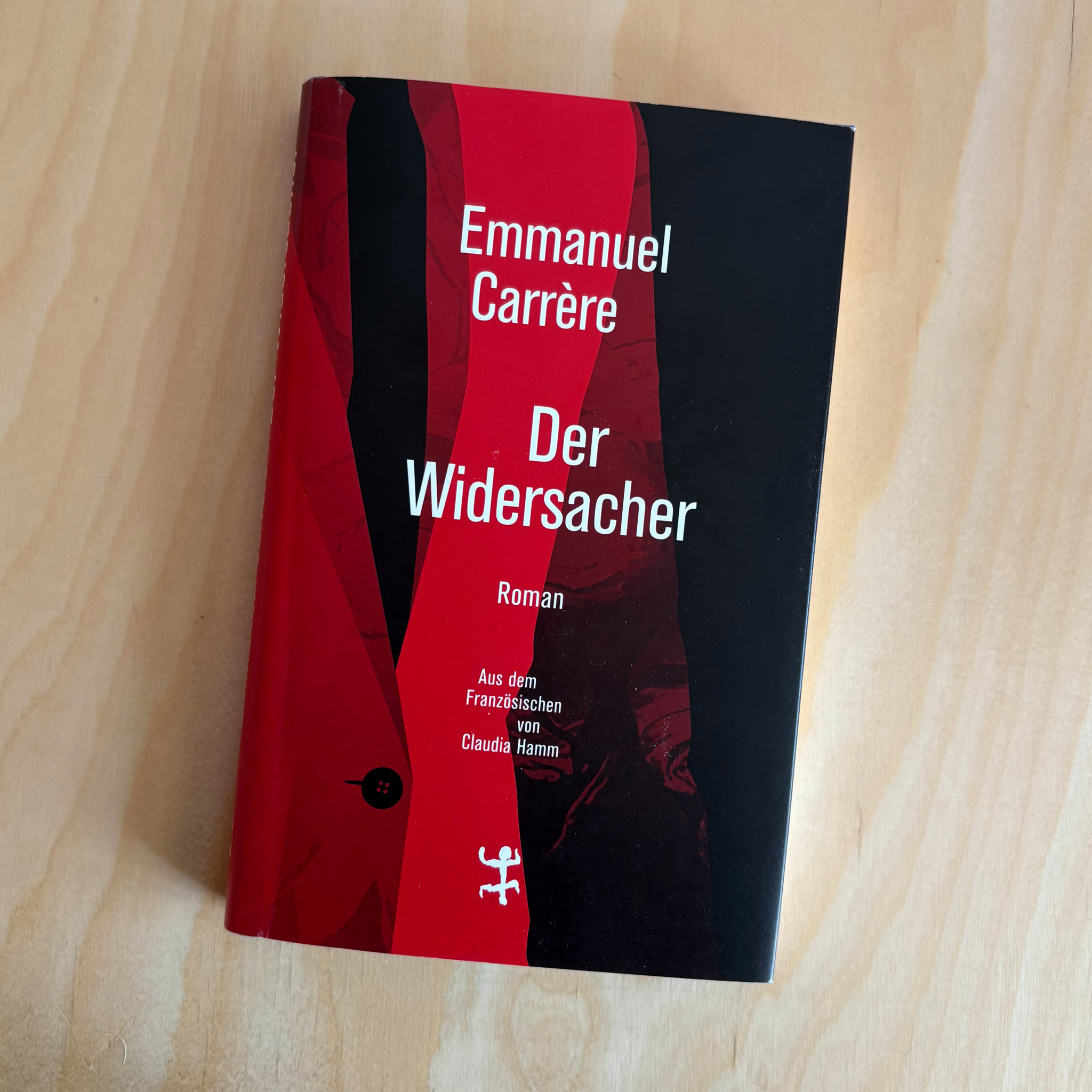 Buchcover von Emmanuel Carrères Roman «Der Widersacher» in Schwarz und Rot, auf einem Holzuntergrund liegend.